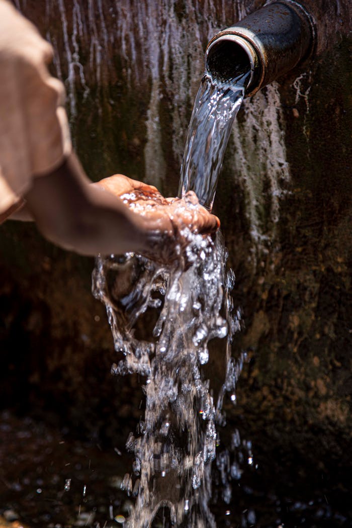 Close-up of hands collecting flowing water from a pipe outdoors.